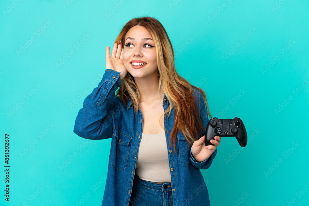 Teenager blonde girl playing with a video game controller over isolated wall listening to something by putting hand on the ear Teenager blonde girl playing with a video game controller over isolated wall listening to something by putting hand on the ear