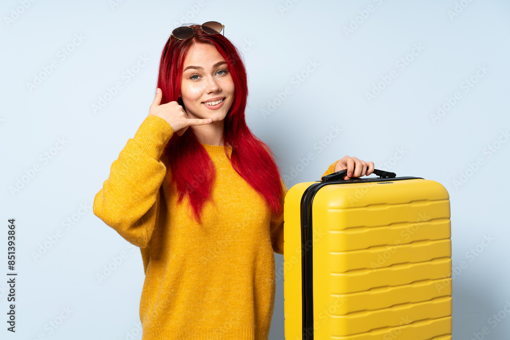Traveler girl holding a suitcase isolated on blue background making phone gesture and pointing front Traveler girl holding a suitcase isolated on blue background making phone gesture and pointing front