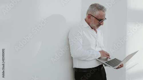 A professional man in a white shirt leans against a white wall while working on a laptop. The minimalist setting and natural light create a clean and focused work environment.