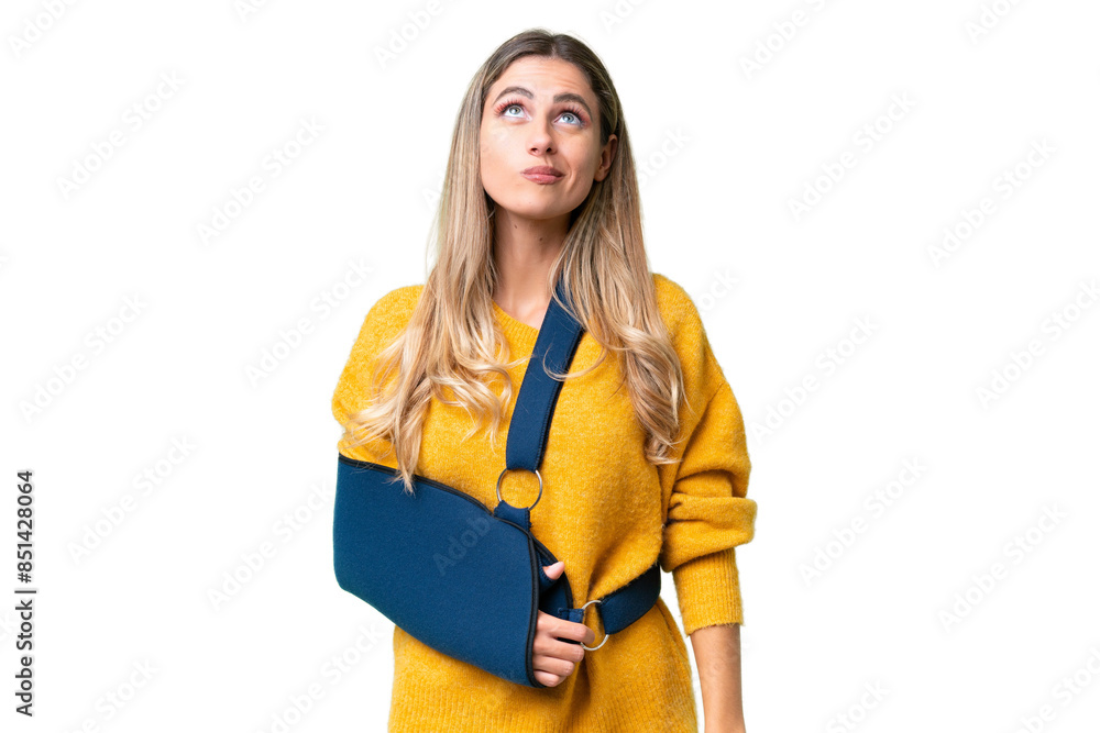 Young Uruguayan woman with broken arm and wearing a sling over isolated background and looking up Young Uruguayan woman with broken arm and wearing a sling over isolated background and looking up