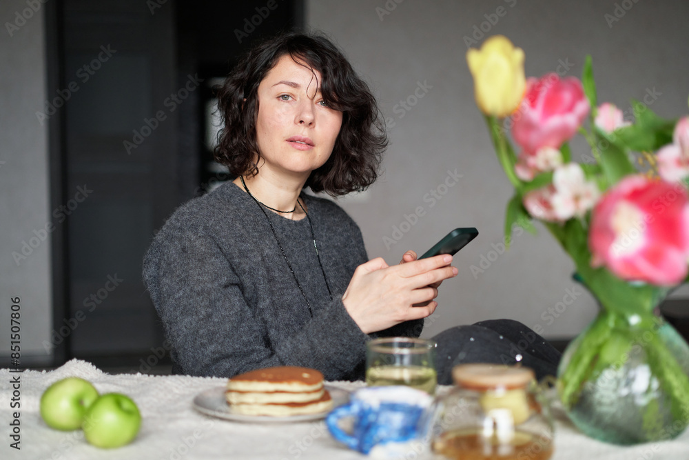 Positive middle age women sitting the the table at home with tea and pancakes. Using smartphone . Beautiful lade looking at the camera, smiling, wearing casual clothing. High quality photo Positive middle age women sitting the the table at home with tea and pancakes. Using smartphone . Beautiful lade looking at the camera, smiling, wearing casual clothing. High quality photo