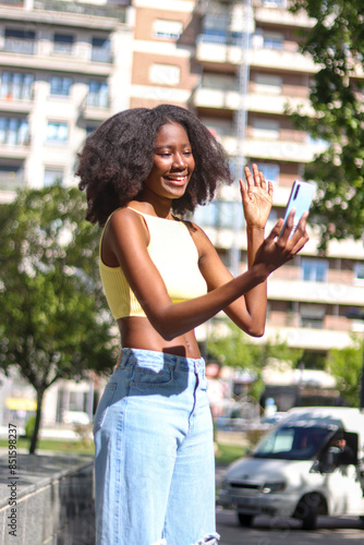 Black woman making a video call with the mobile phone on the street, greeting whoever is calling, vertical image. Concept of social technology