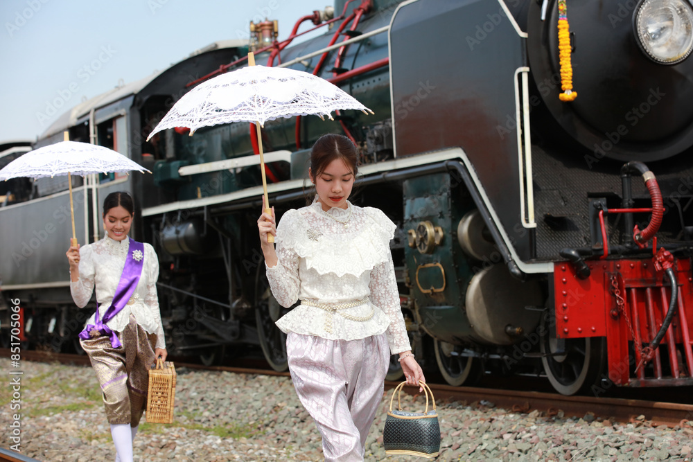 Obraz premium Two Asian women in Thai traditional dress walk with umbrellas across the tracks in front of a steam locomotive at the traditional train station.