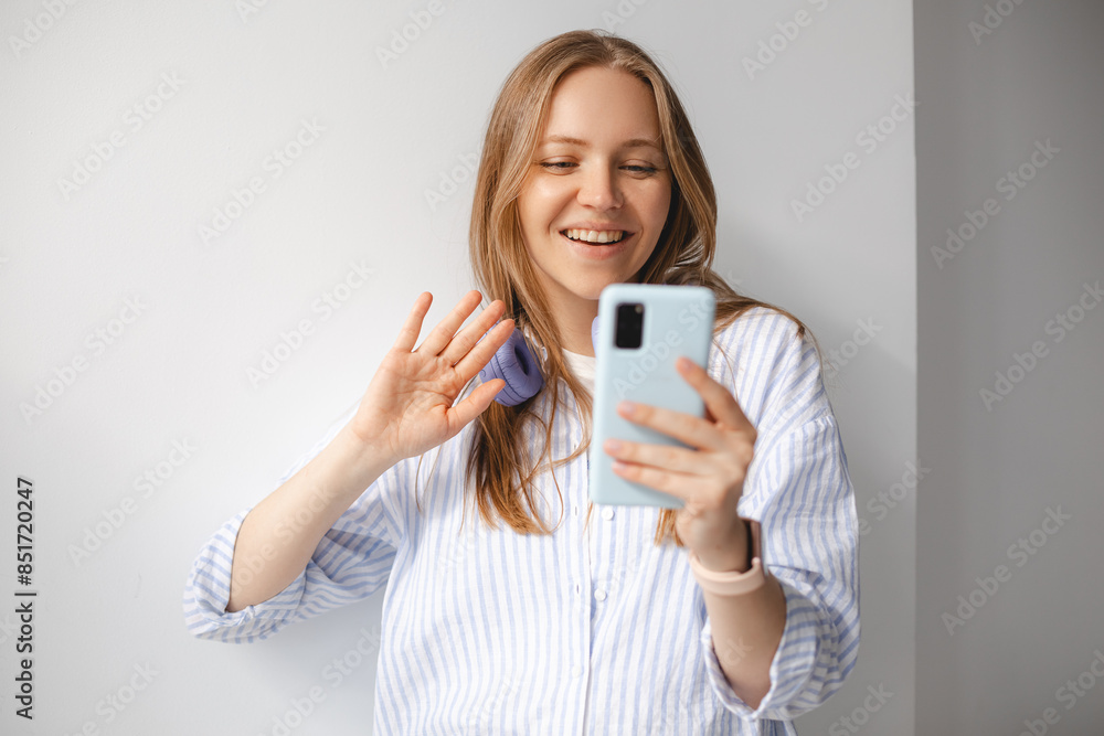 Happy young woman in headphone blogger influencer holding modern smart phone wave hand hello. Smiling girl looking at mobile make video call, shooting vlog taking selfie on white wall background. Happy young woman in headphone blogger influencer holding modern smart phone wave hand hello. Smiling girl looking at mobile make video call, shooting vlog taking selfie on white wall background.
