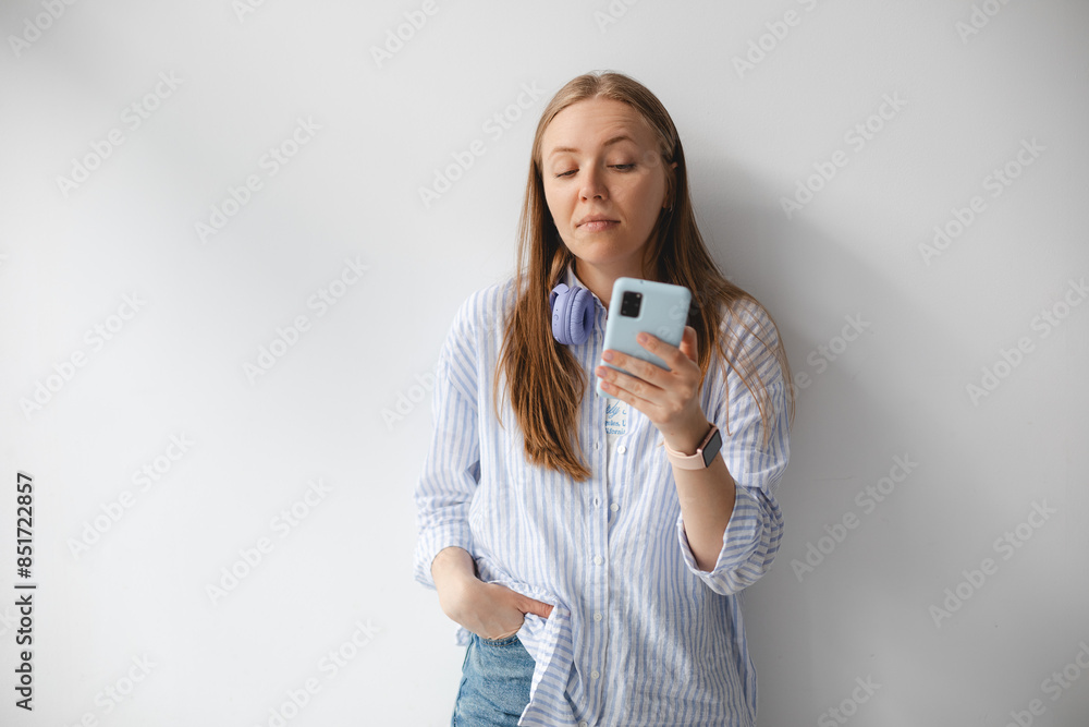 Blonde woman wear shirt, headphones around her neck, her hand in pocket, hold mobile phone, read sms, news. Girl look to phone with boring. Girl feel disinterest, annoyance and tiredness. Blonde woman wear shirt, headphones around her neck, her hand in pocket, hold mobile phone, read sms, news. Girl look to phone with boring. Girl feel disinterest, annoyance and tiredness.