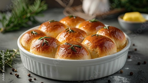 Freshly baked dinner rolls with golden crust in a white ceramic dish, garnished with rosemary and peppercorns on rustic background. photo