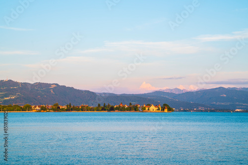 Germany, Warm orange sunset light shining on alps behind water of bodensee in summer with snow covered peaks next to old town of lindau city