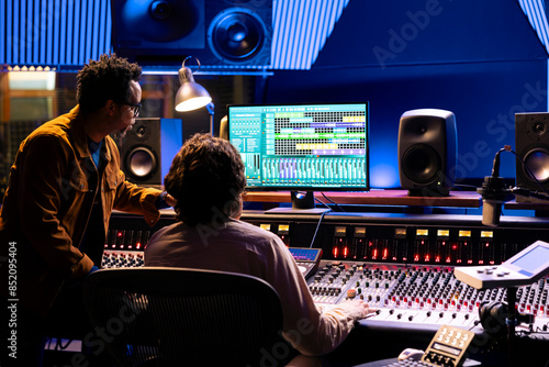 Singer and producer work on soundboard controller at recording studio station. Sound engineer uses digital console for music production, operating on faders and key buttons at control desk.