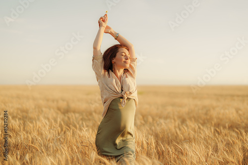 Joyful young woman with arms raised experiencing freedom and happiness in a beautiful golden wheat field at sunset.