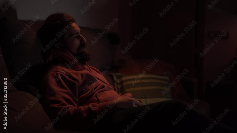 Caucasian man with beard sitting quiet on sofa changing channel with TV remote control. Young casual millennial male smile enjoying movie, documentary or series relaxing comfort at night room home Caucasian man with beard sitting quiet on sofa changing channel with TV remote control. Young casual millennial male smile enjoying movie, documentary or series relaxing comfort at night room home