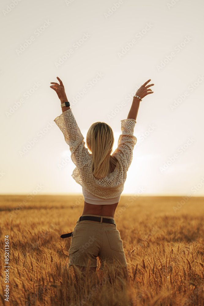Back view of a joyful woman with raised arms feeling free in a beautiful, sunlit wheat field at sunset.