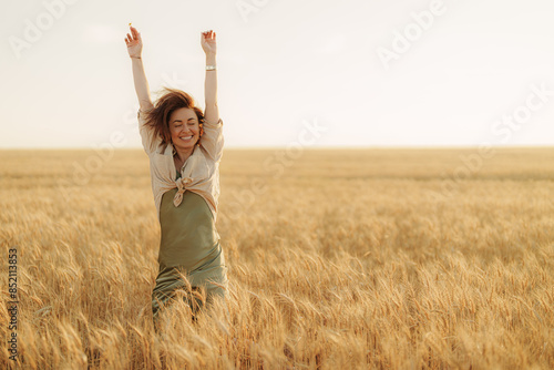 Happy woman with arms raised celebrating freedom and life in a beautiful golden wheat field at sunset, feeling positive and cheerful.