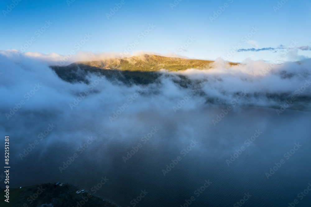 A stunning aerial view of Lake Skarvatnet in Oppdal, Norway, with a mountain ridge shrouded in clouds at dawn. The sun casts a golden glow on the scene A stunning aerial view of Lake Skarvatnet in Oppdal, Norway, with a mountain ridge shrouded in clouds at dawn. The sun casts a golden glow on the scene