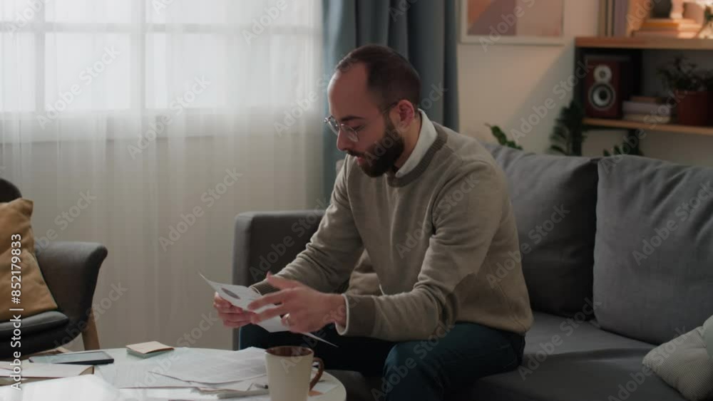 Medium shot of young Middle Eastern man sitting on couch at home, dealing with household expenses, opening envelope, taking out new bill, examining it, clenching fist and swearing Medium shot of young Middle Eastern man sitting on couch at home, dealing with household expenses, opening envelope, taking out new bill, examining it, clenching fist and swearing