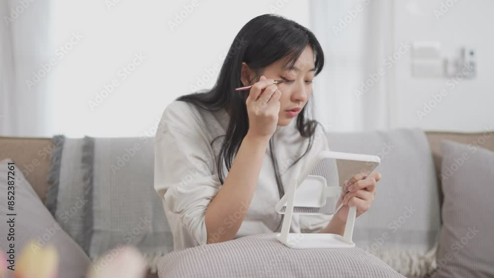 An Asian woman is sitting at a table, applying makeup using a mirror, with beauty products around, in her cozy living room An Asian woman is sitting at a table, applying makeup using a mirror, with beauty products around, in her cozy living room