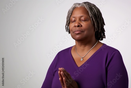 A close-up portrait of an elderly black woman with gray dreadlocks, wearing a purple shirt and a silver necklace, praying with closed eyes against a white background