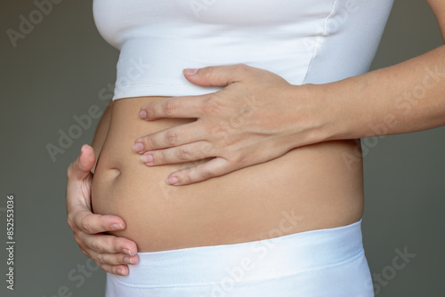 Stomach ache, bloating, flatulence, abdominal distension, colic, digestive disorders. Young woman in profile holding her stomach with hands on a grey background. Overeating, overweight, excess weight