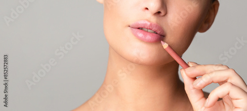 A close-up of a woman face as she carefully applies a pink lip liner to her lips, demonstrating a makeup technique. The background is plain and neutral, cropped, copy space