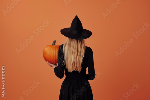 blonde witch wearing a black dress and hat holding a Halloween pumpkin, on a studio smooth orange background with copy space photo