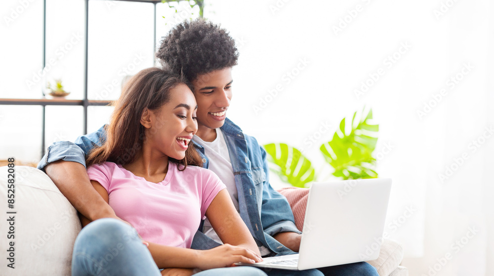 A teen couple is sitting on a couch in their living room. The guy has his arm around the girl, and they are both looking at a laptop. They appear to be relaxed and enjoying each others company.