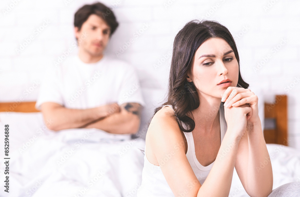 The image shows a woman sitting on a bed, looking sad and lost in thought. She is wearing a white tank top and has her hands clasped together in front of her. The image shows a woman sitting on a bed, looking sad and lost in thought. She is wearing a white tank top and has her hands clasped together in front of her.