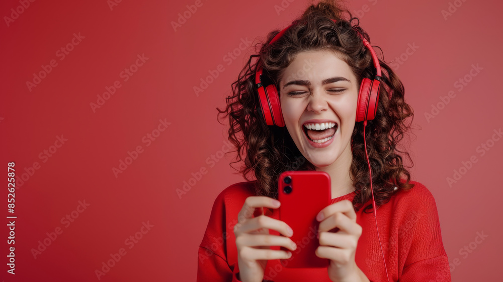 A joyful young woman wearing red headphones, laughing while holding and looking at her red smartphone against a vibrant red background. A joyful young woman wearing red headphones, laughing while holding and looking at her red smartphone against a vibrant red background.