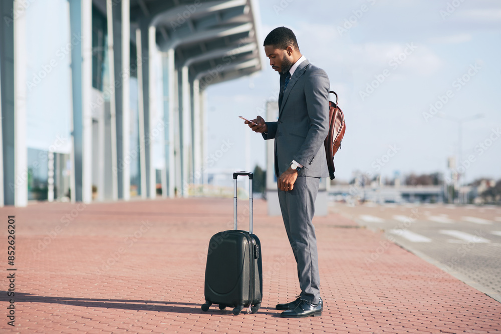 Black businessman in a suit checking his phone while standing with a rolling suitcase outside a modern building. Black businessman in a suit checking his phone while standing with a rolling suitcase outside a modern building.