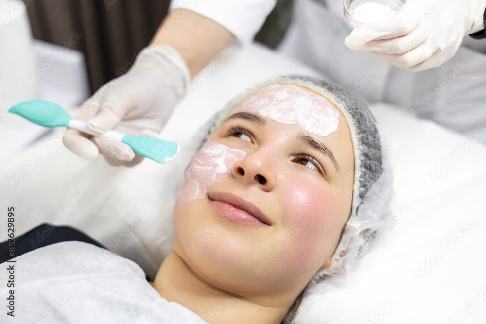Close-up of a young woman in a beauty salon while the dermatologist, beautician, cosmetologist is applying a clay mask with a spatula on her face. Close-up of a young woman in a beauty salon while the dermatologist, beautician, cosmetologist is applying a clay mask with a spatula on her face.