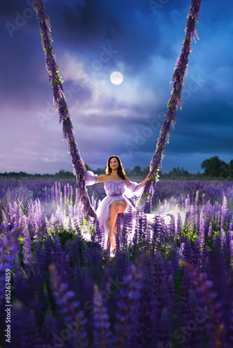 A beautiful young brunette girl in a purple dress sitting on a swing decorated with flowers among a blooming lupine field.