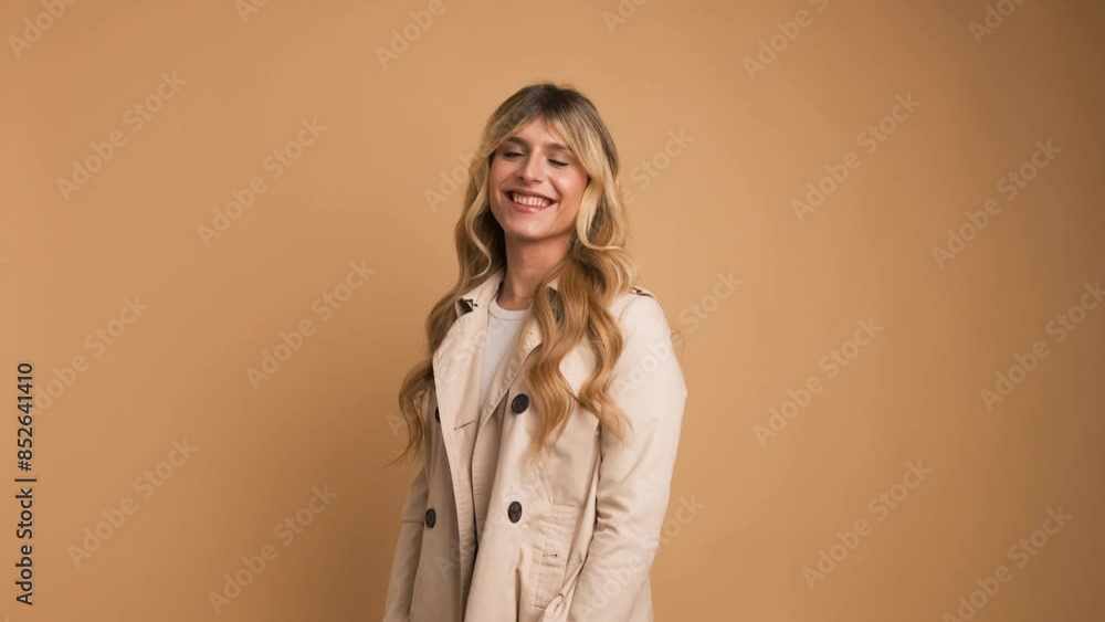 Happy transgender woman in trench coat standing against beige background Happy transgender woman in trench coat standing against beige background