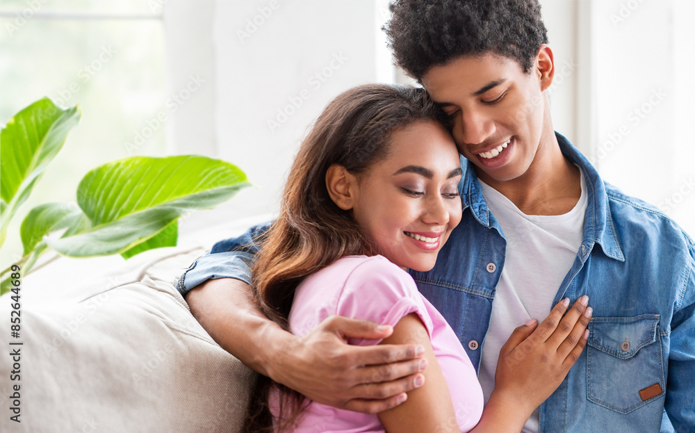 African American teen couple is sitting on a couch indoors. The guy is wearing a blue jean shirt and a white shirt underneath. He is embracing the girl who is wearing a pink shirt African American teen couple is sitting on a couch indoors. The guy is wearing a blue jean shirt and a white shirt underneath. He is embracing the girl who is wearing a pink shirt