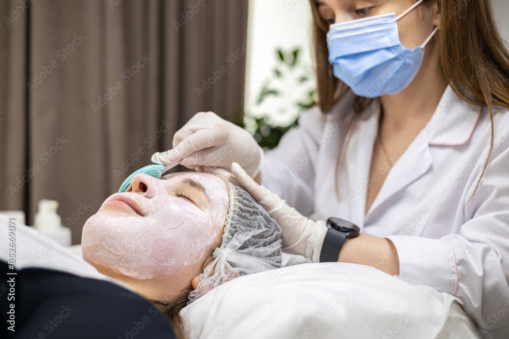 Obraz premium Portrait of a young cosmetologist wearing a surgical mask and white latex gloves, applying a face mask with a spatula on a young woman's face.