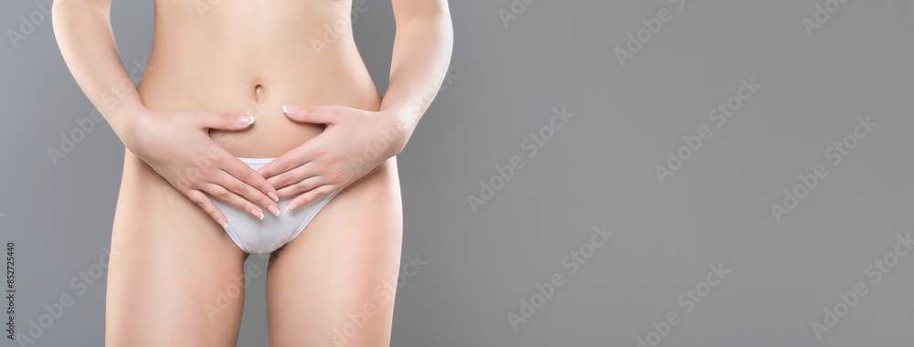 A close-up of a woman gently placing her hands on her lower abdomen, dressed in white underwear, standing against a plain grey backdrop, panorama with copy space A close-up of a woman gently placing her hands on her lower abdomen, dressed in white underwear, standing against a plain grey backdrop, panorama with copy space