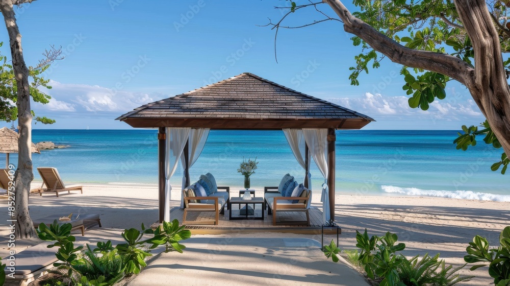 Fototapeta premium A wooden gazebo with white curtains stands on a pristine beach, offering a relaxing view of the turquoise ocean. The beach is covered in white sand, and there are lounge chairs and greenery around the
