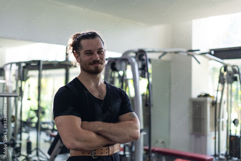 A man with a beard and a ponytail is posing for a picture in a gym. He is wearing a black shirt and a belt A man with a beard and a ponytail is posing for a picture in a gym. He is wearing a black shirt and a belt