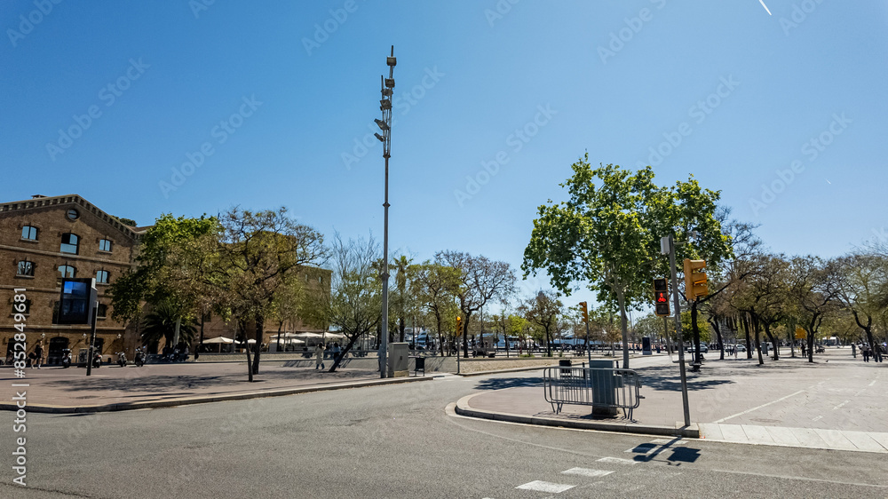 Obraz premium Sunny urban square with lush green trees, traffic lights, and a surveillance camera pole on a clear day, ideal for themes of city planning and public safety