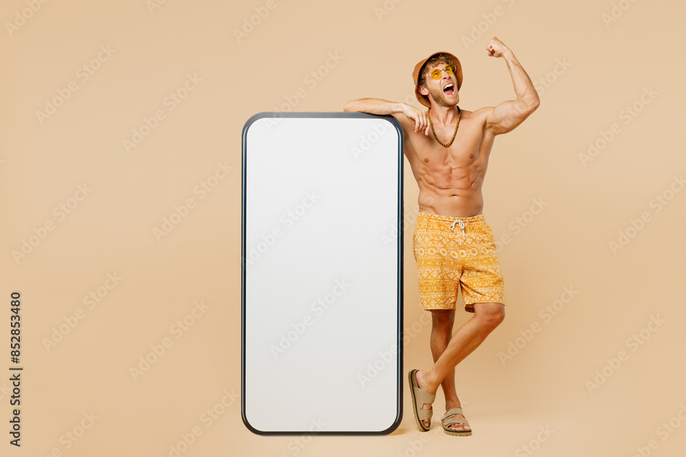 Full body young man wear yellow shorts swimsuit hat relax rest near hotel pool big huge blank screen mobile cell phone do winner gesture isolated on plain beige background. Summer sea sun tan concept. Full body young man wear yellow shorts swimsuit hat relax rest near hotel pool big huge blank screen mobile cell phone do winner gesture isolated on plain beige background. Summer sea sun tan concept.