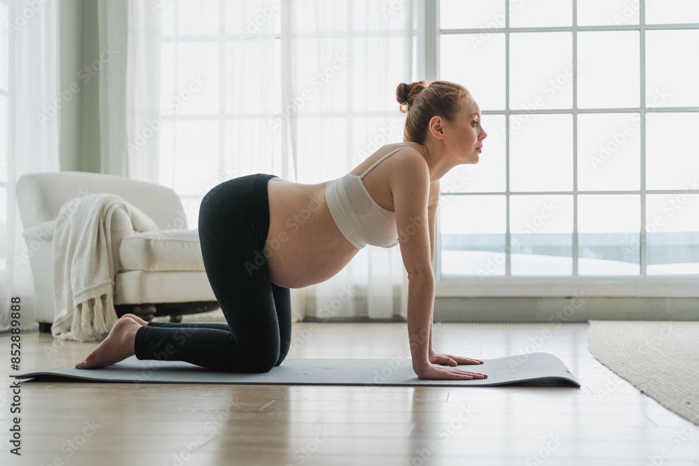 Fototapeta premium Pregnancy yoga fitness workout training. Pregnant woman practicing yoga at home. Pregnant girl doing sports exercise on yoga mat indoor. Woman with big belly have fitness training practicing asana