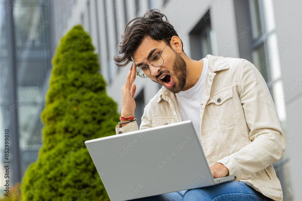 Displeased Indian man use laptop typing browsing working, loses becoming surprised sudden lottery results bad news fortune loss game fail computer virus. Arabian sad guy sitting on street city bench Displeased Indian man use laptop typing browsing working, loses becoming surprised sudden lottery results bad news fortune loss game fail computer virus. Arabian sad guy sitting on street city bench