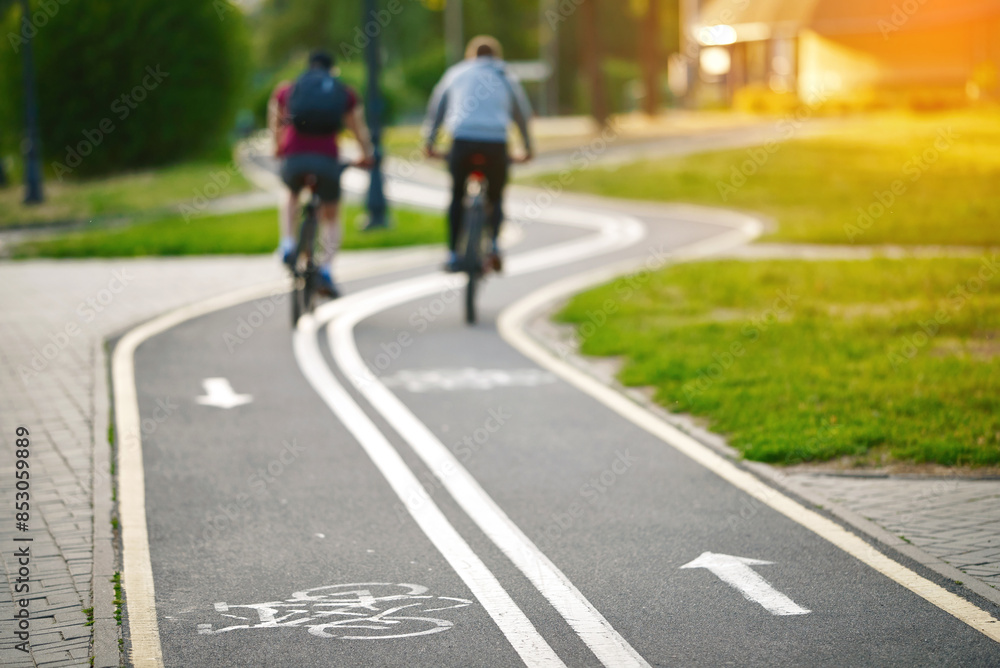 Curvy bike lane with markings and men cycling in the background, recreational and fitness benefits of cycling. Bicycle lane in park. Bicyclists riding. Selective focus. Curvy bike lane with markings and men cycling in the background, recreational and fitness benefits of cycling. Bicycle lane in park. Bicyclists riding. Selective focus.