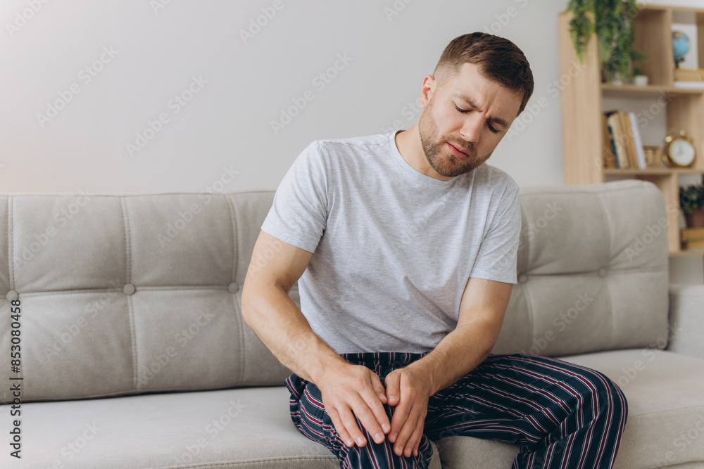 Unhappy man sitting on sofa at home and touching hurt knee with grimace of pain and suffering. Young guy with rheumatism, arthritis or osteoarthritis feeling intense severe pain in knee joint Unhappy man sitting on sofa at home and touching hurt knee with grimace of pain and suffering. Young guy with rheumatism, arthritis or osteoarthritis feeling intense severe pain in knee joint