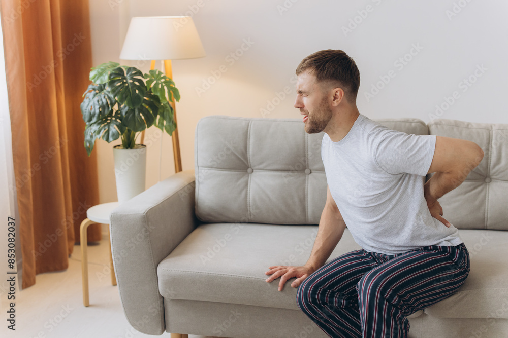 Tired bearded man suffering from back pain sitting on couch in living room at home. Male massages painful lower back muscles. Caucasian guy exhausted overworked in apartment. Tired bearded man suffering from back pain sitting on couch in living room at home. Male massages painful lower back muscles. Caucasian guy exhausted overworked in apartment.