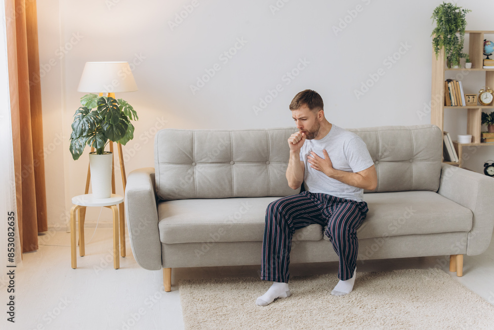 Sick man holding his chest in pain while coughing in the living room. Sore throat and cough, man with lung pain at home, health problems concept Sick man holding his chest in pain while coughing in the living room. Sore throat and cough, man with lung pain at home, health problems concept