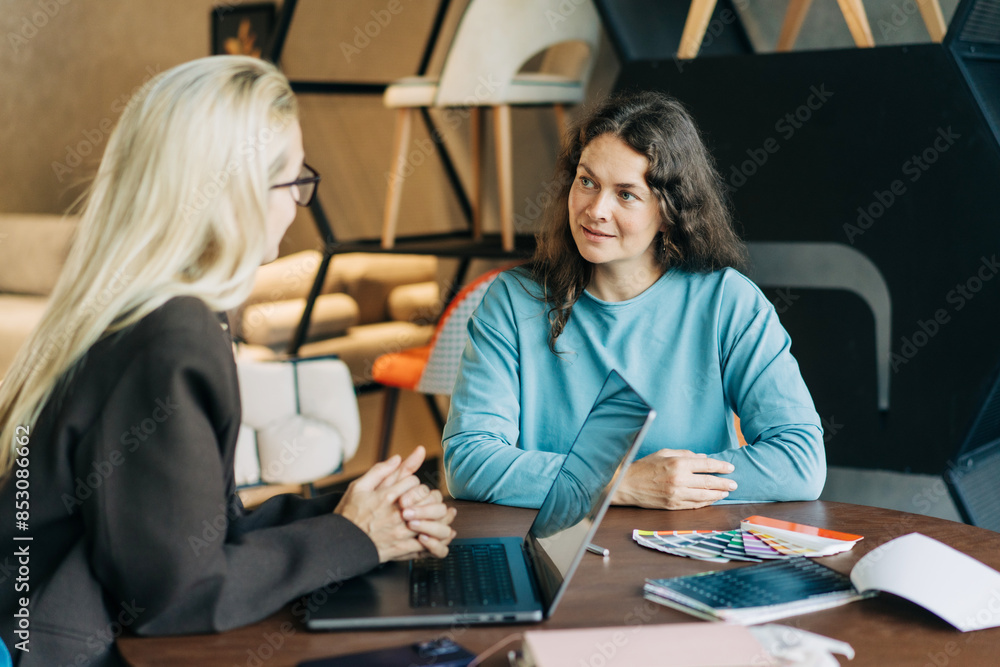 Two business women are discussing while sitting at an office table in a furniture showroom. Two business women are discussing while sitting at an office table in a furniture showroom.