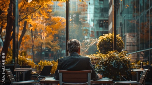 A man in a suit sits alone at an outdoor cafe table, surrounded by autumn foliage and modern architecture with a laptop on the table.