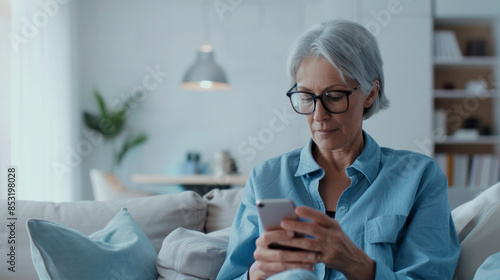 Serious, grey-haired senior woman relaxing at home, keenly typing a message on her smartphone with glasses on, on her cozy living room sofa. photo