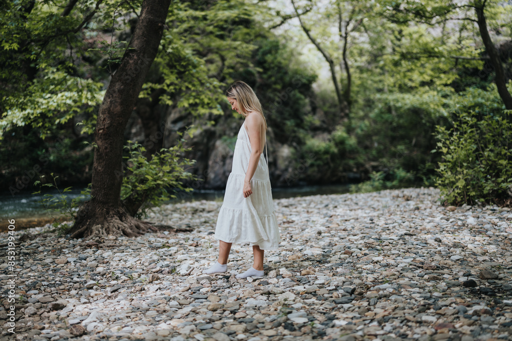 A woman in a white dress is seen walking peacefully in a lush green forest, surrounded by trees and a serene river. A woman in a white dress is seen walking peacefully in a lush green forest, surrounded by trees and a serene river.