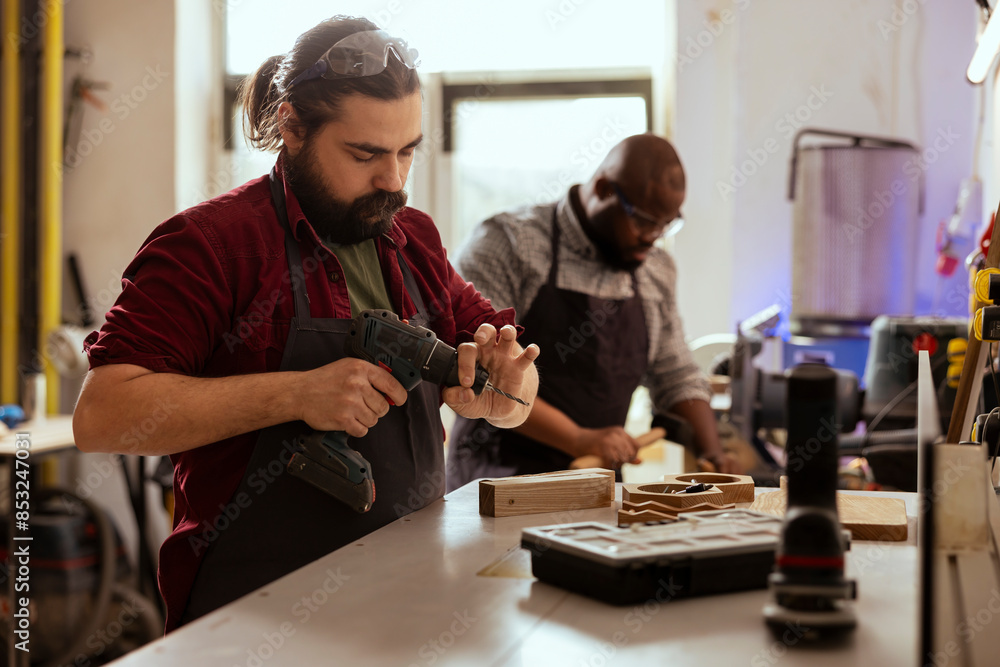 Fototapeta premium Woodworker next to african american coworker changing power drill head. Carpenter working with BIPOC colleague, preparing to insert screws into wooden surfaces with electric tool
