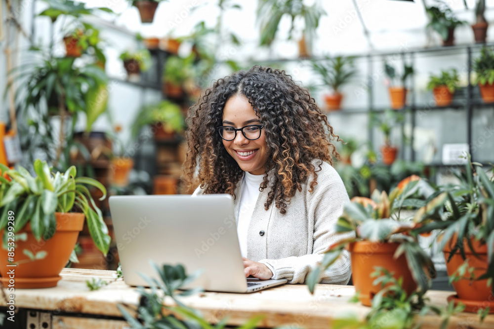 A woman with curly hair and glasses sits at a table in a greenhouse, smiling as she works on a laptop A woman with curly hair and glasses sits at a table in a greenhouse, smiling as she works on a laptop