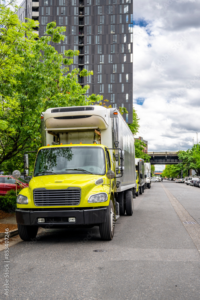 Fototapeta premium Yellow day cab middle duty rig semi truck with refrigerated box trailer standing on the urban city street in front of another trucks